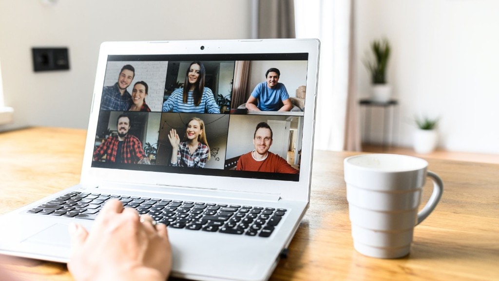 Laptop showing a six-person video call grid on a wooden desk with a coffee mug nearby